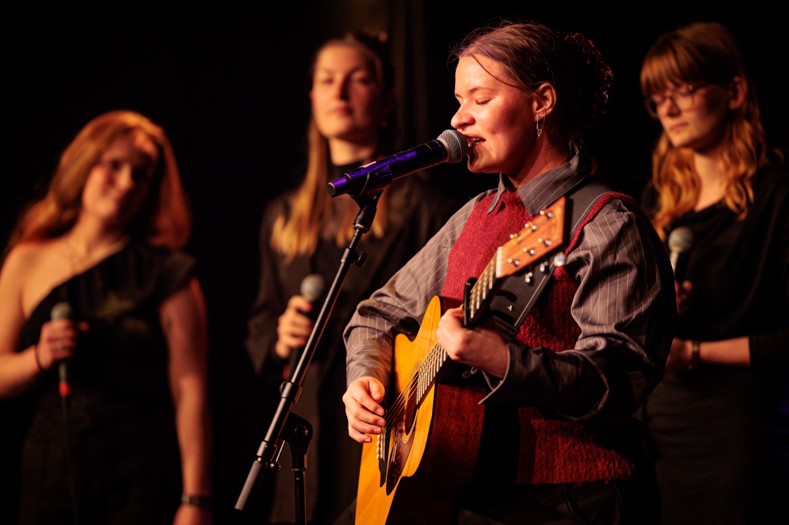 Maria-Louise Lundqvist synger og spiller akustisk guitar på en scene. Bag hende står tre korsangere.